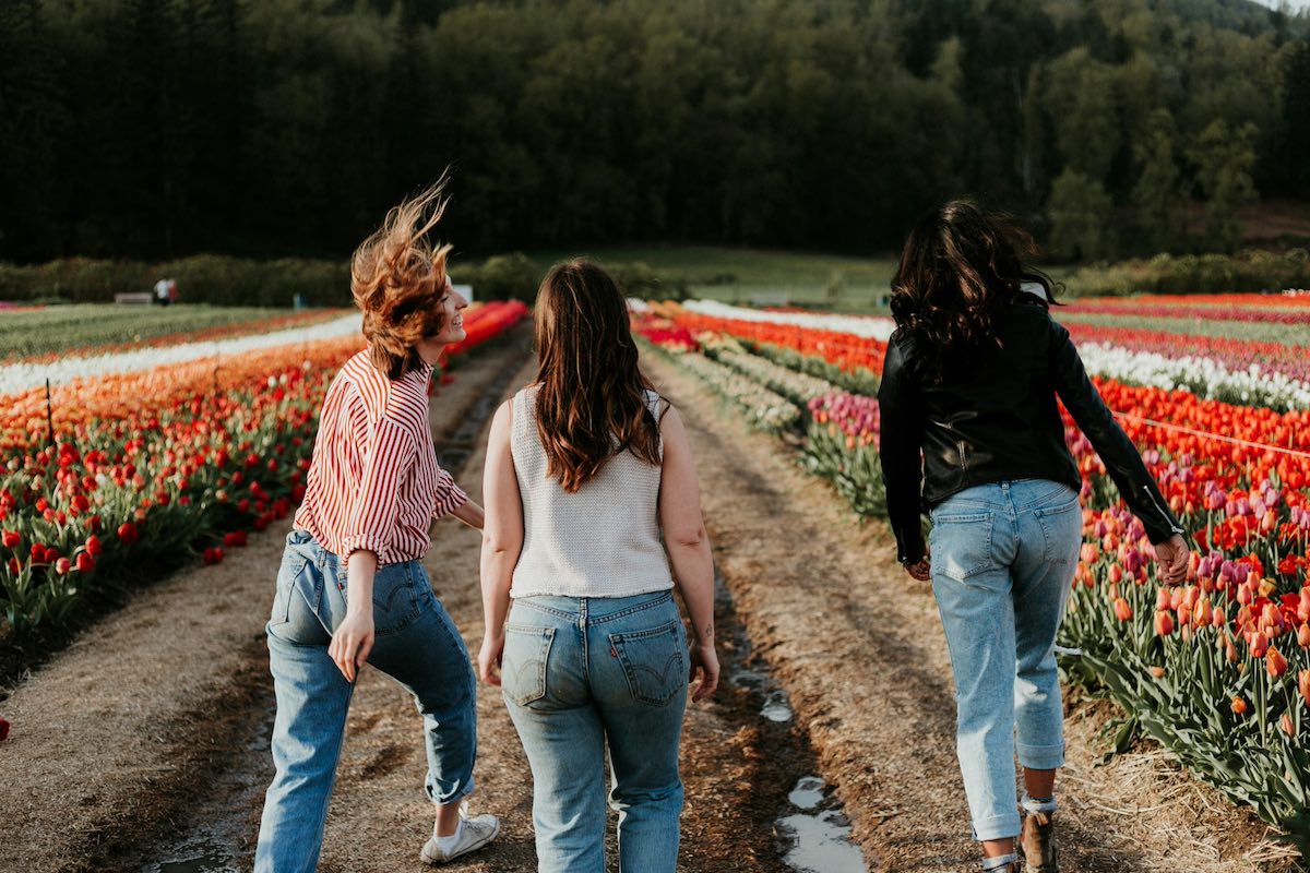 Tre ragazze single riprese da dietro camminano felici in un campo pieno di fiori